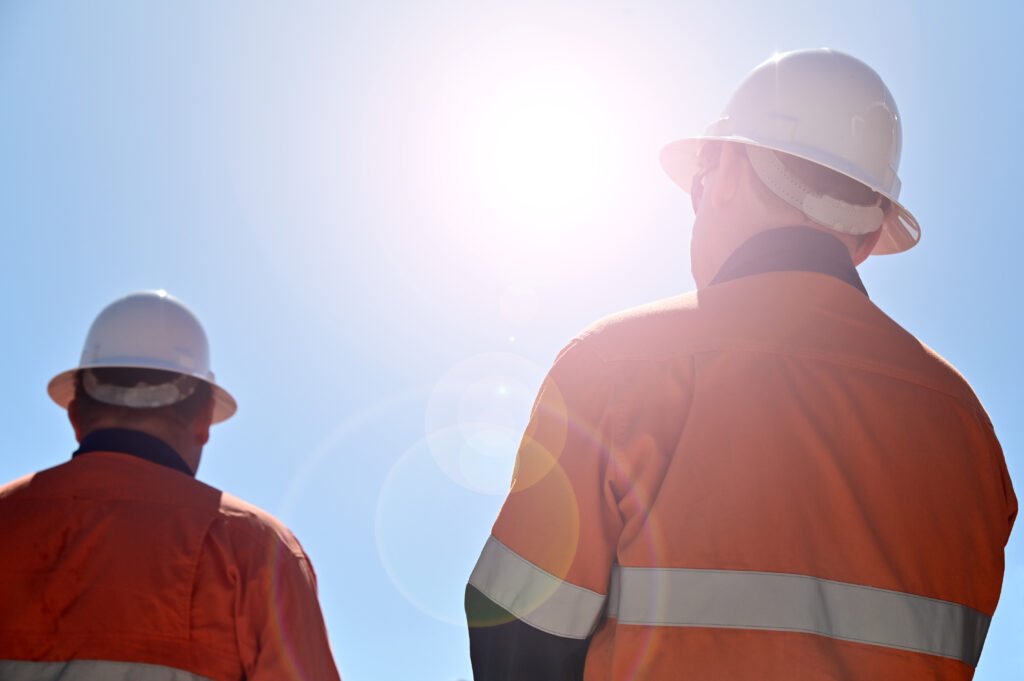 Two construction workers wearing white hard hats and orange jackets standing outside in the sun with a blue sky.