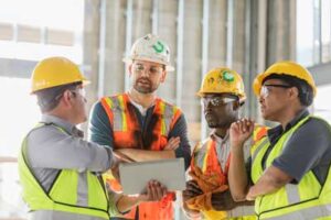 four construction workers wearing vests and hardhats looking at a laptop