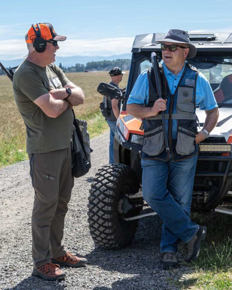 two men talking, one holding a rifle and leaning against a four wheeler