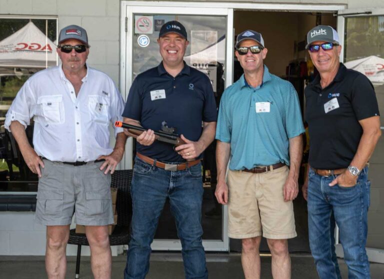 four men smiling, one holding a rifle